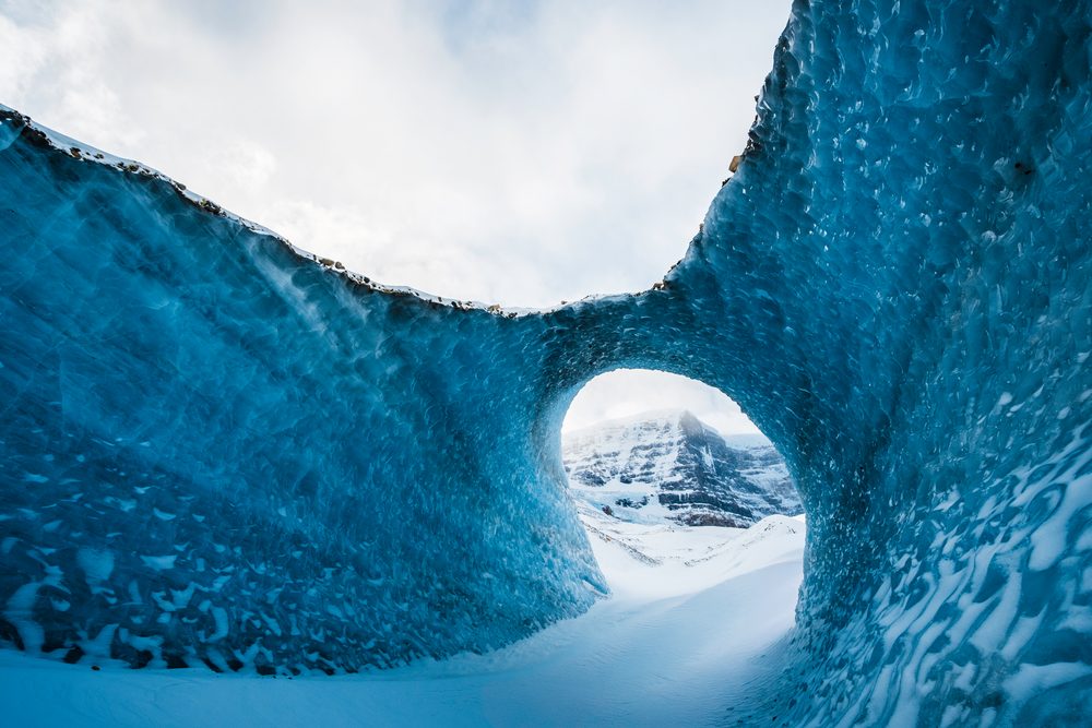 A wave shaped arch of blue ice framing a small round opening, with a snowy peak beyond.