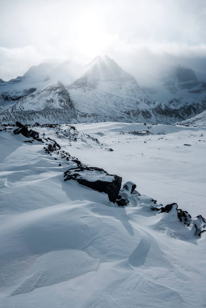 A dark ridge of rock cutting through snow drifts, with sun rays breaking through cloud above the peaks.