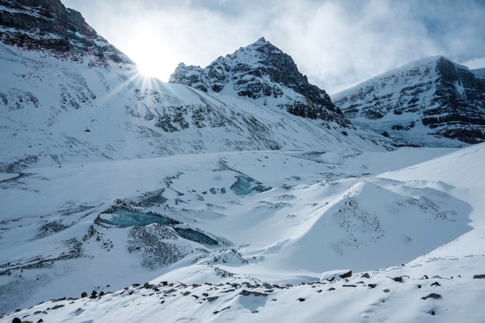 Low sun bursting over a snowy ridge, with blue pools of ice scattered across the glacier.