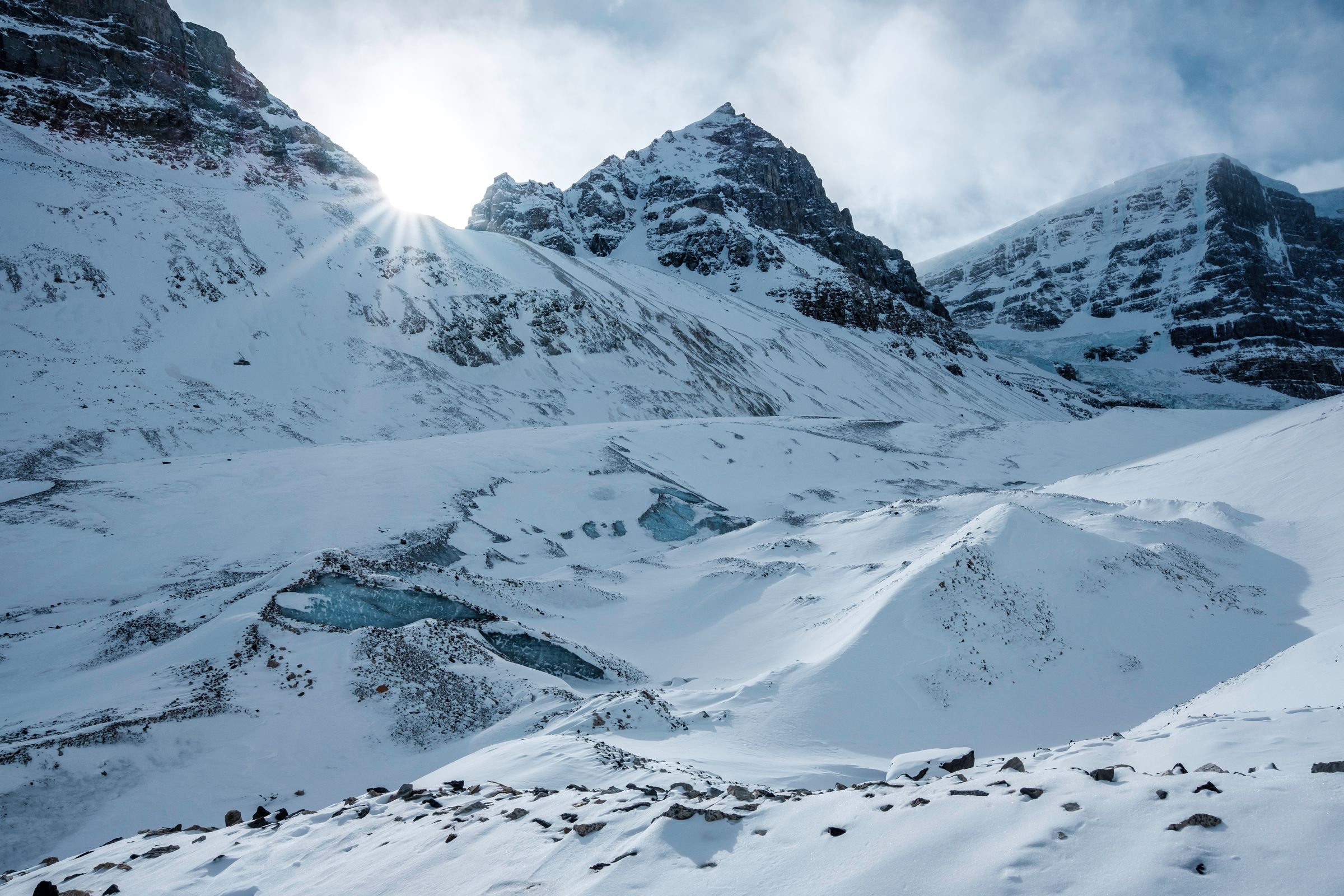 Low sun bursting over a snowy ridge, with blue pools of ice scattered across the glacier.