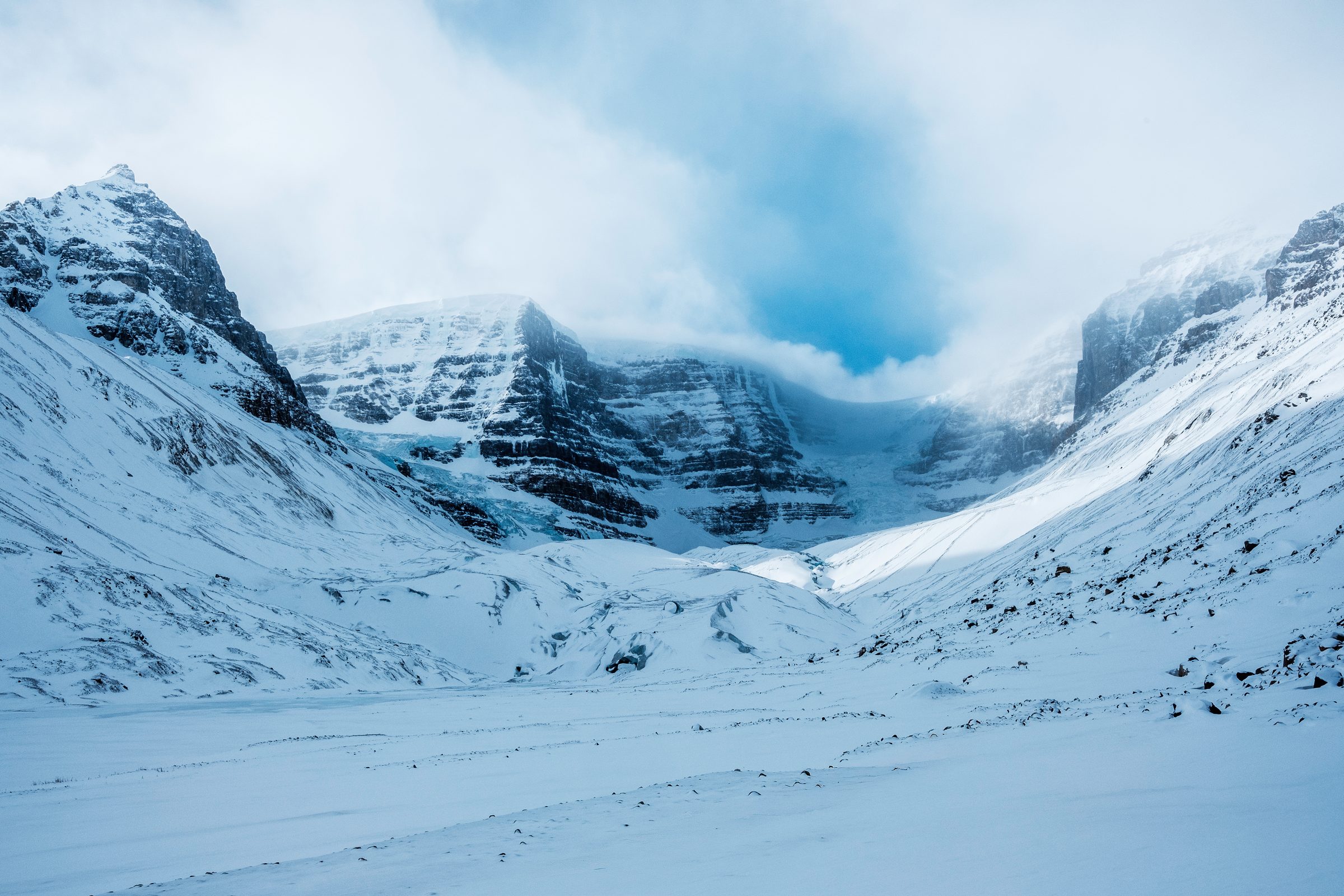 A wide snow covered valley with steep rock walls and a band of cloud breaking over the peaks.