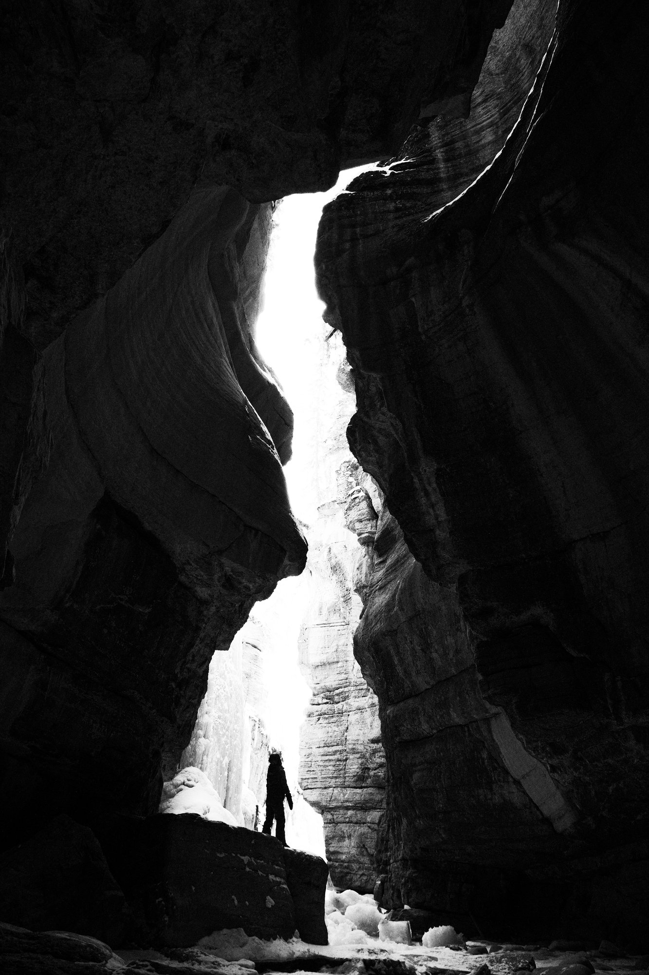Black and white narrow canyon of curved rock walls with bright light pouring through the gap above a lone figure standing on a rock.