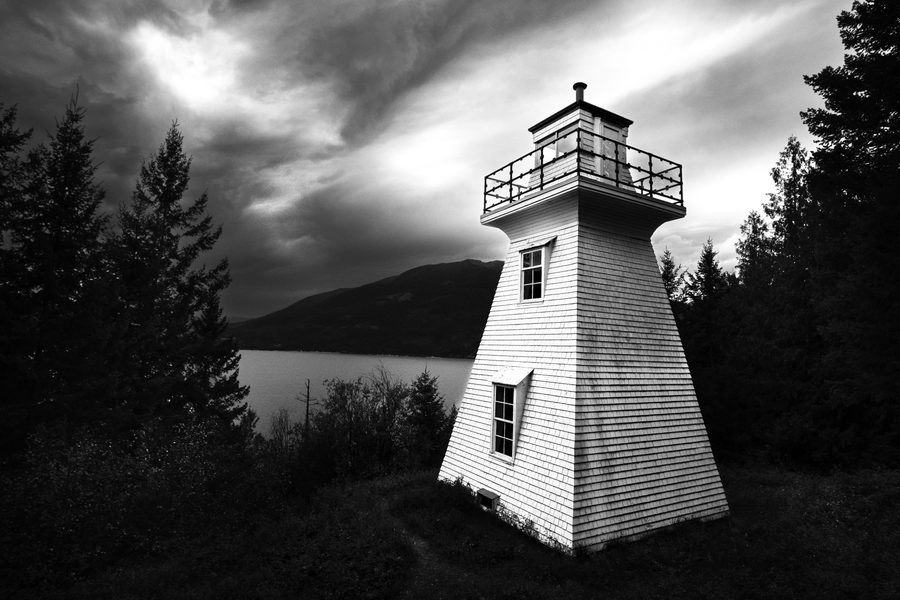 Black and white lighthouse on a rocky point under a stormy sky, dark trees around it and mountains across the water.