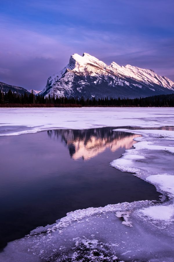 A snowy peak catching first light above a frozen lake, purple ice and open water holding its reflection.