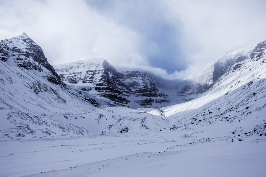 A wide snow covered valley with steep rock walls and a band of cloud breaking over the peaks.