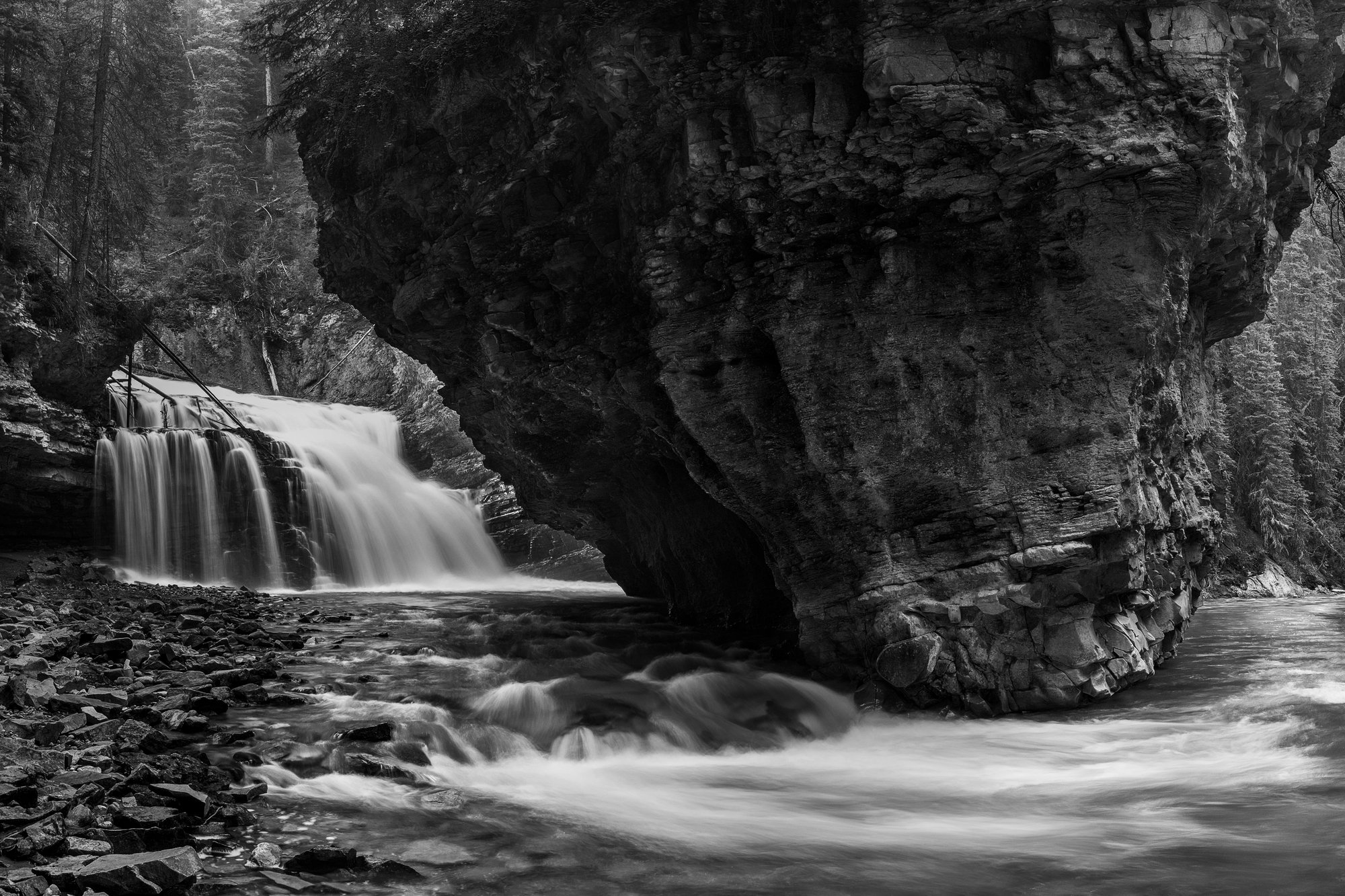 Black and white long exposure of a waterfall pouring into a river beside a huge overhanging block of rock in a forest canyon.