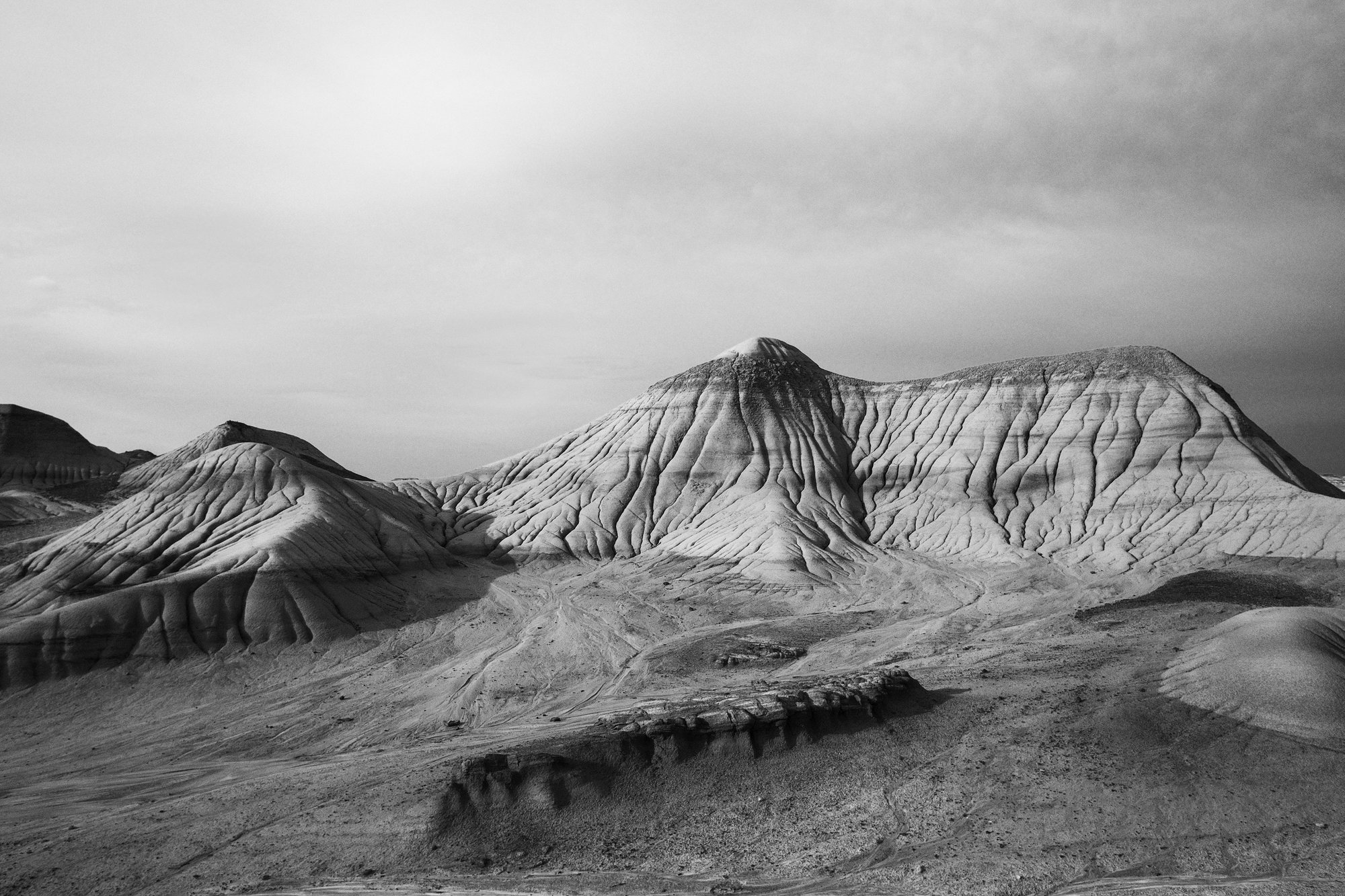 Black and white badlands hills with deep folded ridges and channels under a heavy sky.