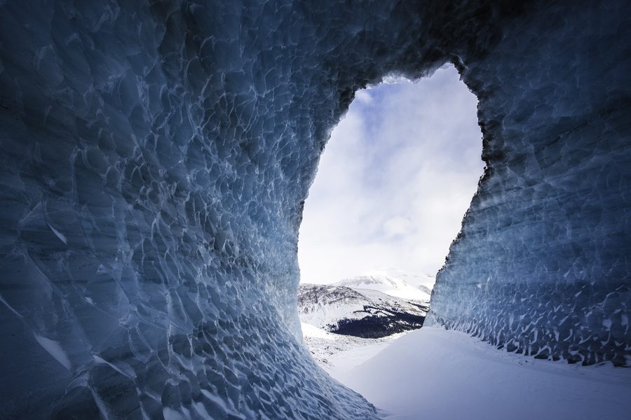 A tall narrow opening in blue ice, with snow outside and a sliver of sky.