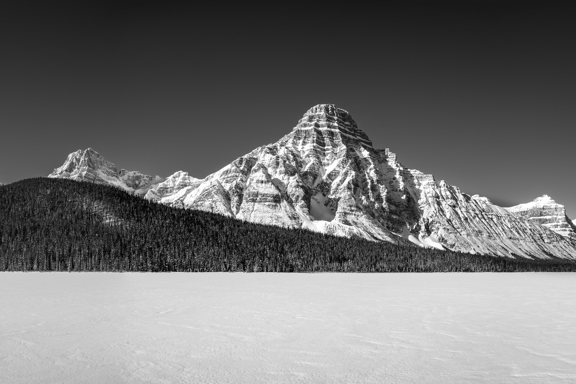 Black and white snowy peak rising above a dark forested ridge and a wide frozen lake under a dark sky.