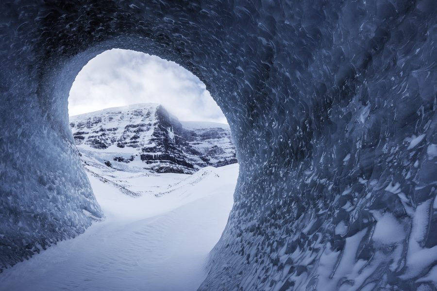 Looking out through a round opening in a wind carved ice tunnel to a snowy peak under cloud.
